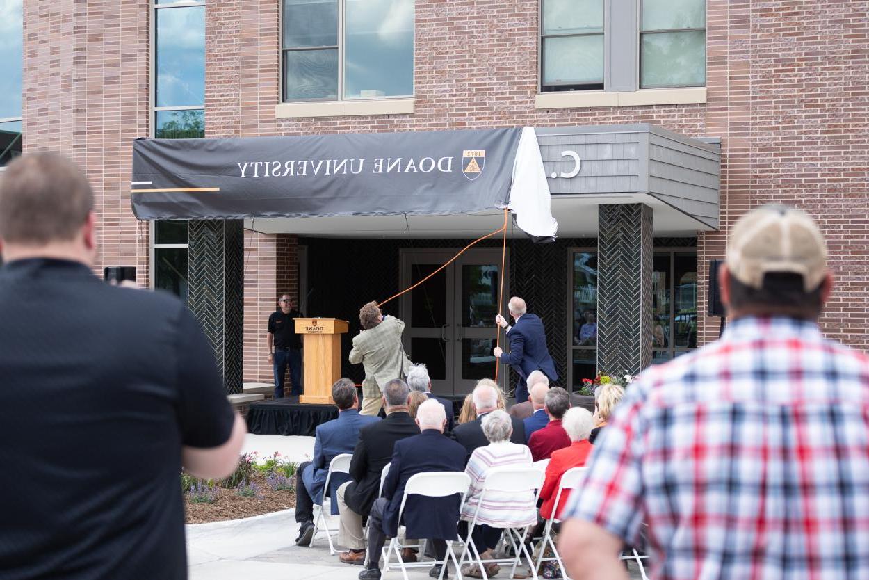 Two men in suits use ropes to remove a banner and reveal the name of the building in the background. They are surrounded by a seated audience with people standing toward the back. 